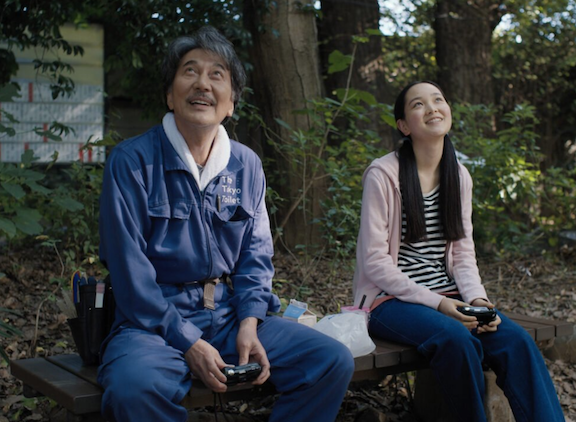 A middle aged Japanese man and a young Japanese woman sit on a bench beneath a tree, looking up with pleasant expressions on their faces.